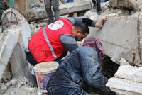 Zwei Männer - einer von ihnen trägt eine Weste des Syrisch Roten Halbmondes - suchen in Trümmern eines zusammengestürzten Hauses nach Überlebenden.