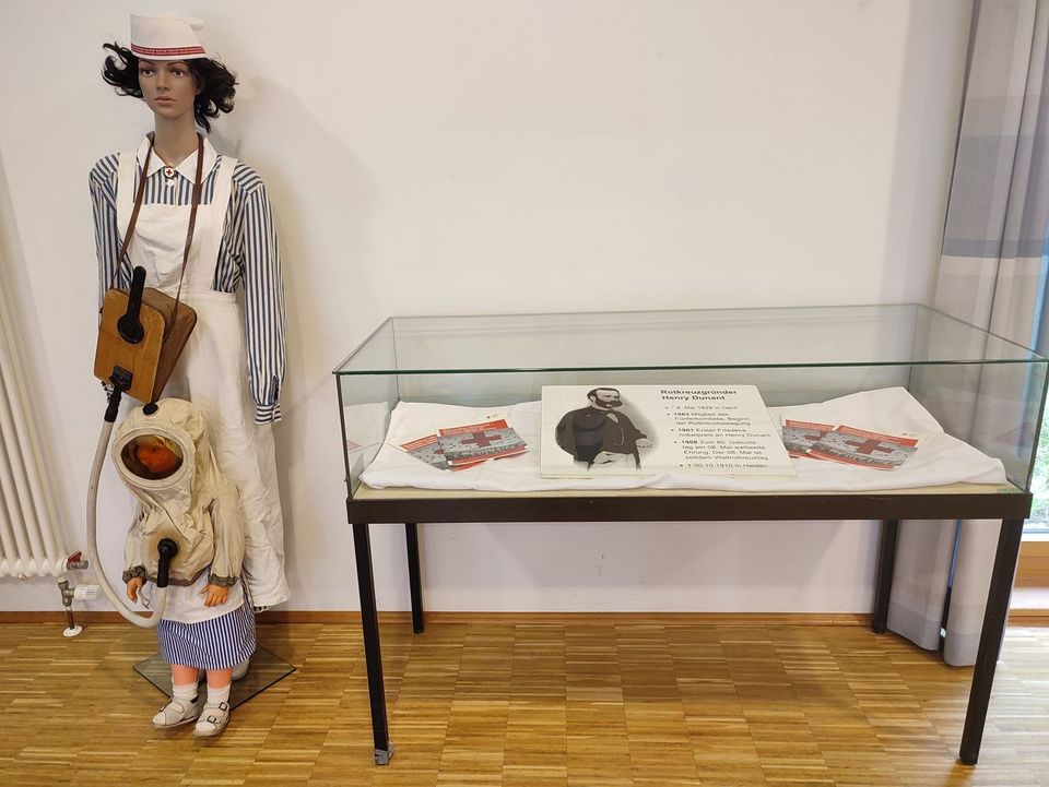 DRK-Wanderausstellung In einer Vitrine liegt eine Tafel mit Infos zu Henry Dunant. Links daneben steht eine Puppe in Rotkreuzschwestern-Kluft.