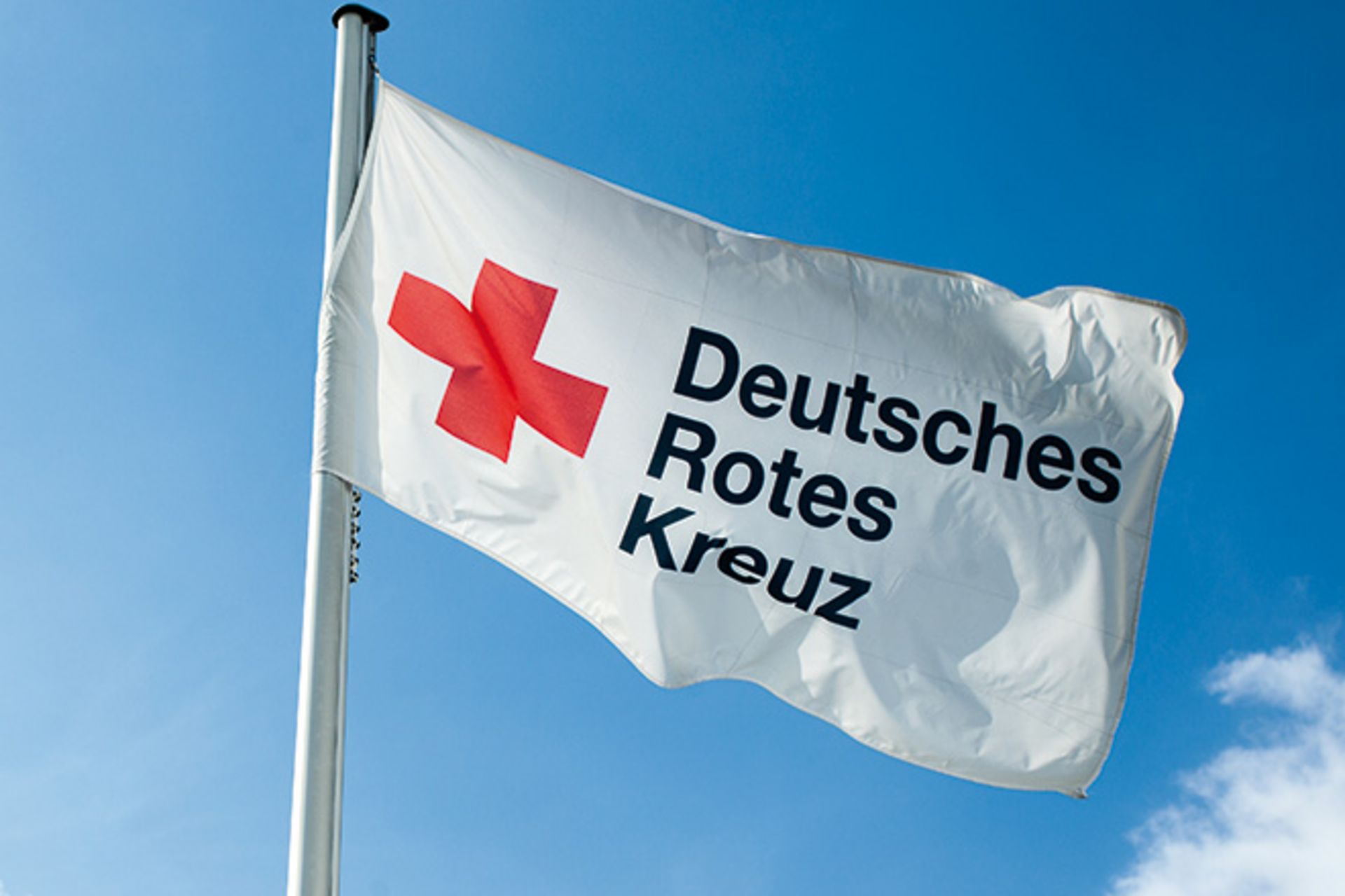 Im Wind weht eine weiße Flagge. Sie zeigt ein rotes Kreuz. Daneben steht in schwarzen Buchstaben: Deutsches Rotes Kreuz.