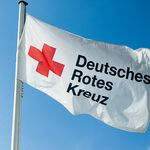 Im Wind weht eine weiße Flagge. Sie zeigt ein rotes Kreuz. Daneben steht in schwarzen Buchstaben: Deutsches Rotes Kreuz.