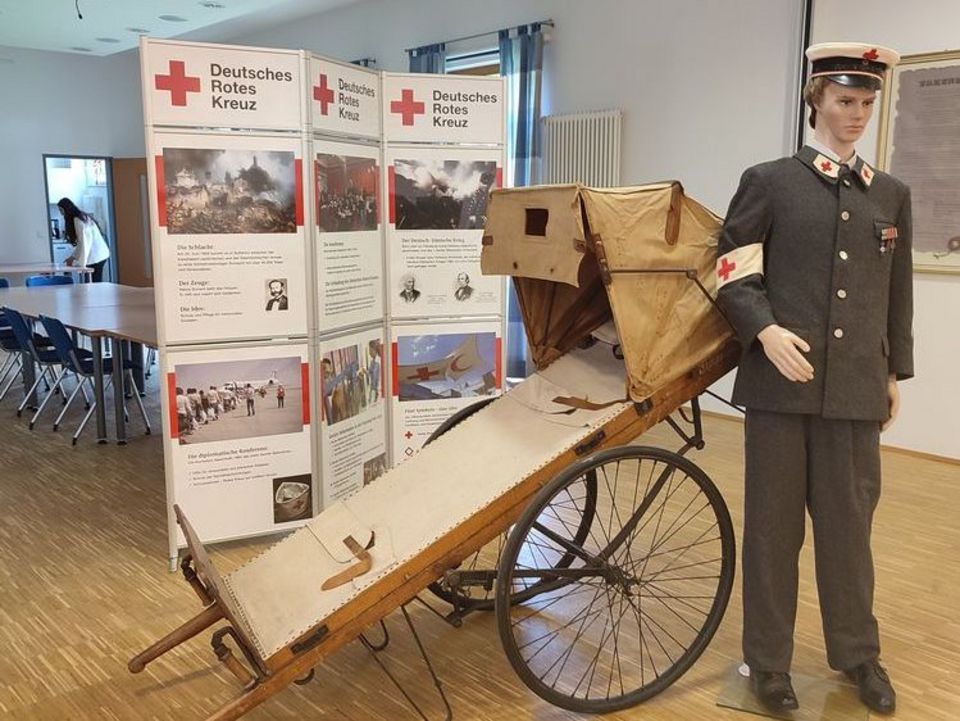 DRK-Wanderausstellung In einem Saal stehen große DRK-Stellwände mit Infos zur Geschichte des humanitären Völkerrechts. Davor steht eine alte hölzerne Patientenliege mit Rädern. Daneben trägt eine Puppe eine graue Uniform mit zahlreichen Rotkreuz-Symbolen.