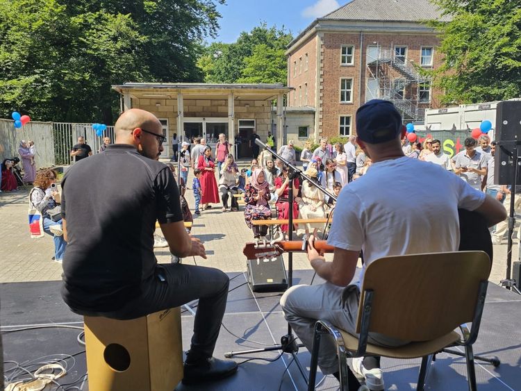 Sommerfest 2025 Blick von hinten auf zwei Personen, die auf einer Bühne Cajon und Gitarre spielen. Im Hintergrund erkennt man das Publikum: Rund 50 Leute.