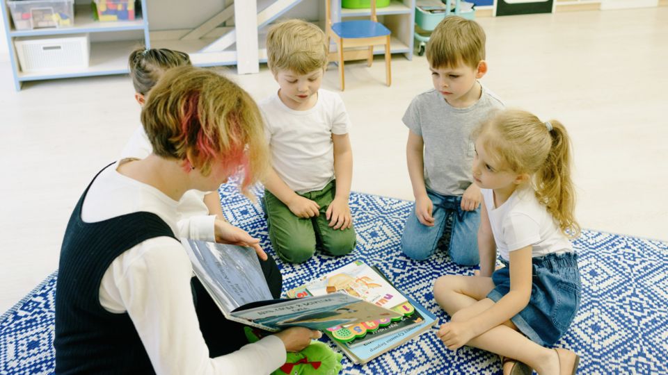 Vier Kinder sitzen auf einem Teppich um eine Erzieherin herum, die aus einem Buch vorliest.